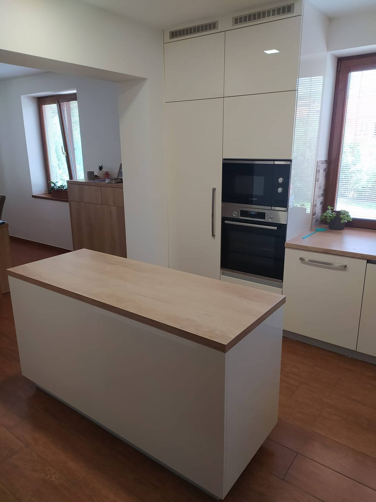 Weiße Küche mit hohem Kasten am Fenster und Insel in der Mitte