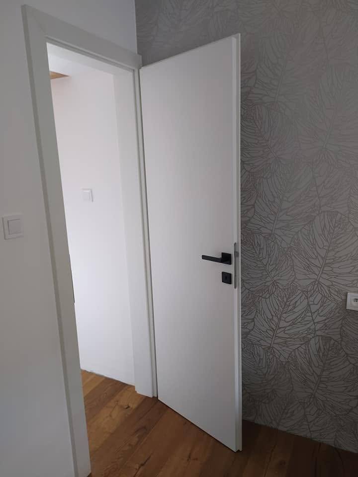 Offene weiße Tür mit schwarzem Griff, die in ein Zimmer mit Holzboden führt.
