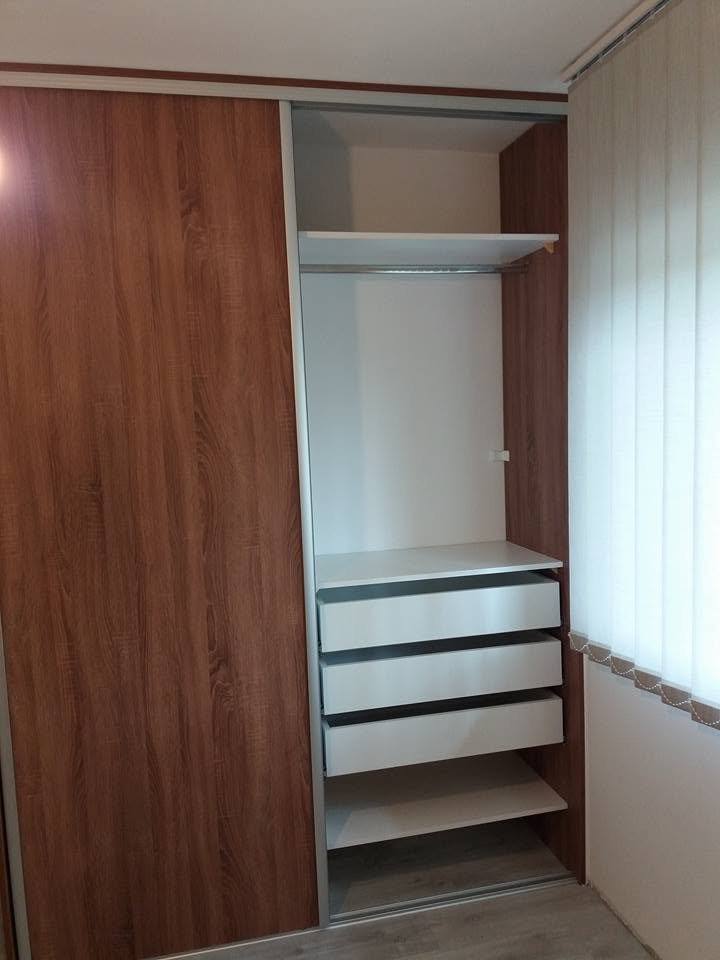 Kleiderkasten in Nussbaum-Dekor mit offenem Innenraum und weißen Laden am Fenster.
