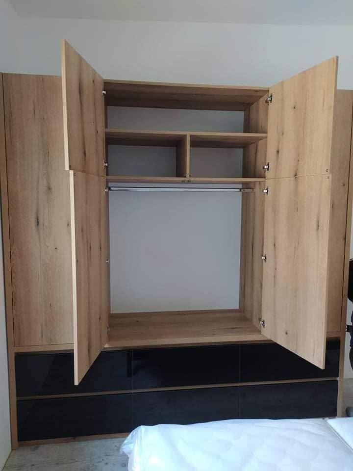 Offener Kleiderkasten in Eichendekor mit Kleidung an der Stange und schwarzen Laden.