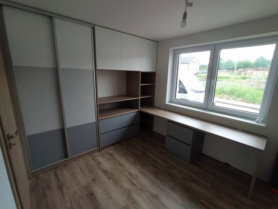 Zimmer mit Eckschreibtisch am Fenster und grau-weißem Schwebetürenkasten