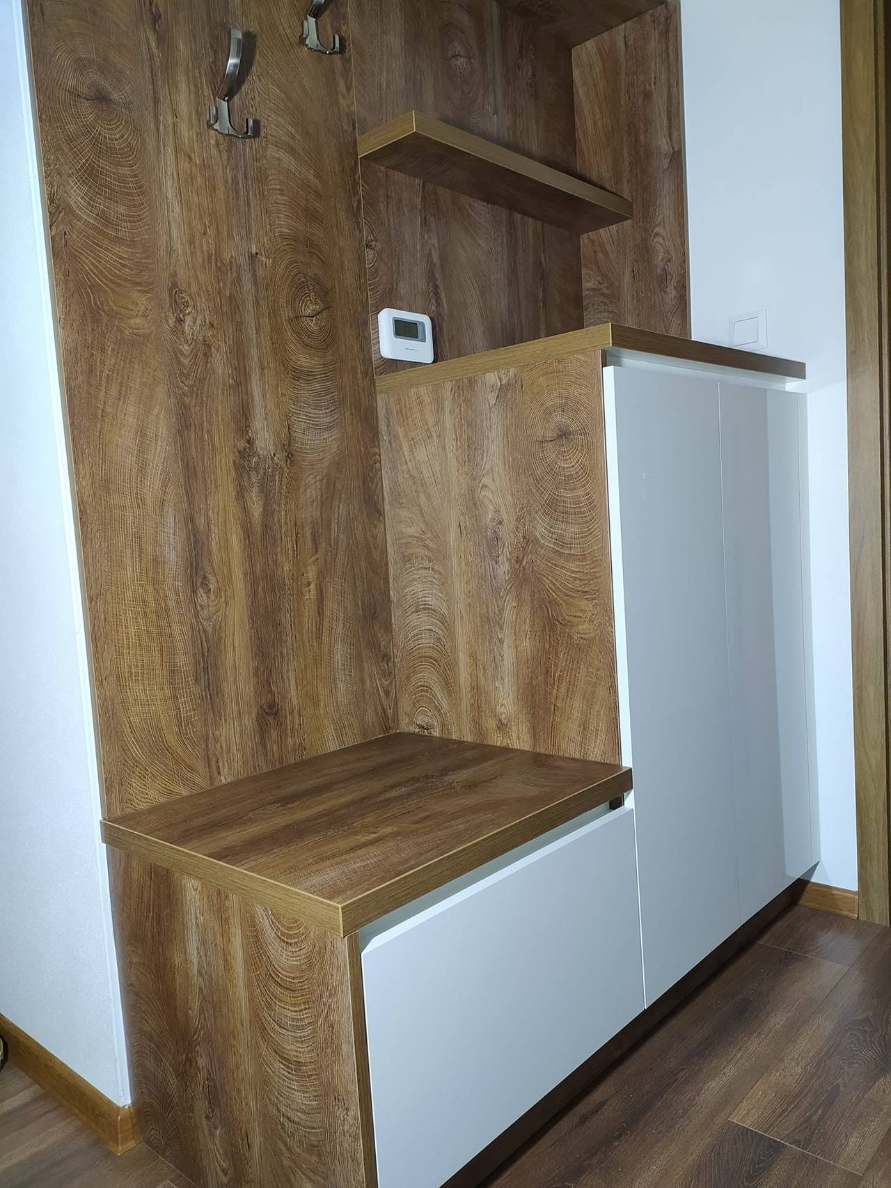 Tmavý drevodekorový panel s háčikmi nad bielou skrinkou a lavicou